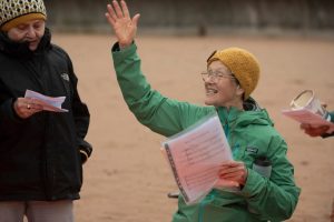 A woman wearing a yellow hat conducting singing on the beach