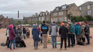 A group of people all wrapped up in coats and hats singing round a fire on the beach